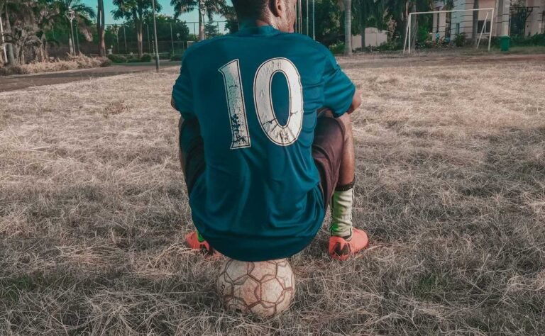 Chico con la camiseta número 10 sentado sobre una pelota en un potrero